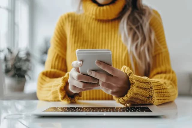 Businesswoman typing on laptop while holding smartphone in bright office with natural light