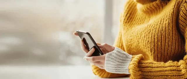 Young woman in yellow sweater using smartphone at office desk, close finger tapping screen