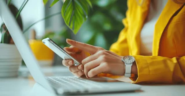 Woman’s hands tapping a smartphone beside laptop keyboard in a modern office desk